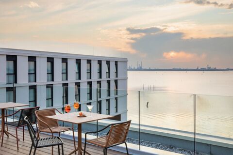 Outdoor seating area with drinks and snacks on table overlooking water and modern building at B&B Hotel Venezia Laguna