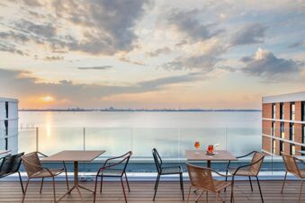 View from a terrace at B&B Hotel Venezia Laguna showing tables, chairs, glasses with drinks, glass railing, and water at sunset