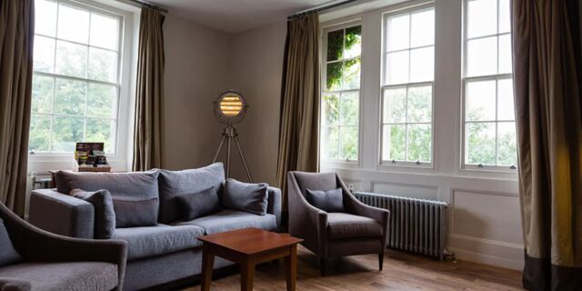 Hotel du Vin Tunbridge Wells living room with gray sofa, two armchairs, wooden table, large windows with beige curtains, and a tripod floor lamp
