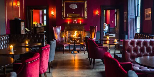 Cozy lounge area with red and gray chairs around tables, a fireplace with candles, and dark wood floors at Hotel du Vin Tunbridge Wells