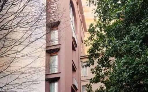 Pink exterior facade of The Corner Duomo Hotel surrounded by leafless and green trees under a warm sky