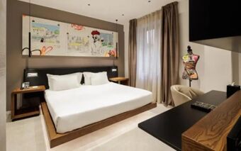 A modern hotel room at The Corner Duomo Hotel with a large bed, bedside tables, abstract wall art, and a mannequin near the window