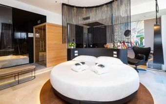 Round bed with white linens and bathrobes in a modern room with black curtain divider and colorful wall art at The Corner Duomo Hotel