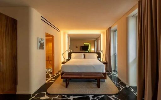 A spacious hotel room at Hotel Calimala with a large bed, wooden bench, nightstands with lamps, and large windows with curtains