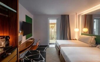 Hotel Calimala guest room with two beds, balcony door, TV mounted on wall, desk, chair, and modern lighting fixtures