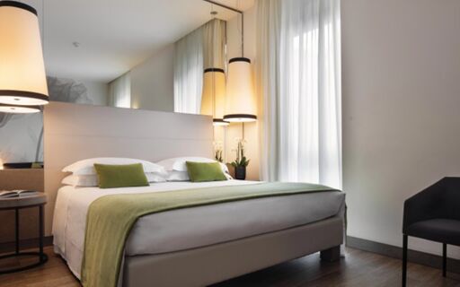 A modern hotel room at Starhotels E.c.ho. with a large bed, white linens, green pillows, hanging lamps, and sheer curtains