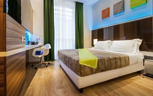 Hotel degli Arcimboldi room with a large bed, brown quilt, green runner, white chair, wooden furniture, and floor-to-ceiling window
