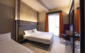 A modern hotel room at Joy 124 Milano Hotel with two beds, a large window with orange curtains, and a desk with a chair
