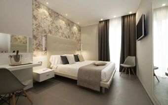 Modern hotel room at Hotel H21 with a double bed, decorative pillows, floral wallpaper, and a wall-mounted TV near the window