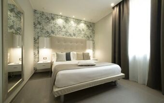 A modern hotel room at Hotel H21 with a large bed, patterned wallpaper, bedside tables with lamps, curtains, and a full-length mirror