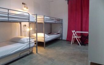 Room with two metal bunk beds, white bedding, wall lamps, a white table with a chair, and a red curtain at Hostel Colours