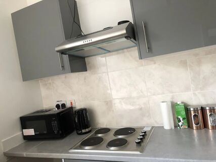 A kitchen counter at Leman House with a black microwave, black kettle, stovetop with four burners, paper towels, milk carton, and two copper canisters