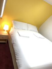 Double bed with white linens and pillows in a corner room with yellow and white walls and a bedside table lamp at Leman House