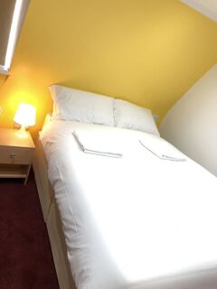 Double bed with white linens and pillows in a corner room with yellow and white walls and a bedside table lamp at Leman House