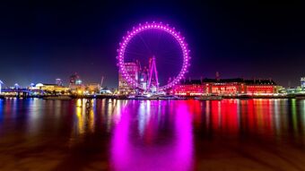 Night view of the illuminated London Eye in pink and surrounding buildings reflecting in the river near Leman House