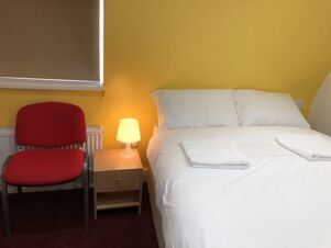 White bed with two pillows and towels beside a wooden nightstand with a lit lamp and a red chair in Leman House room