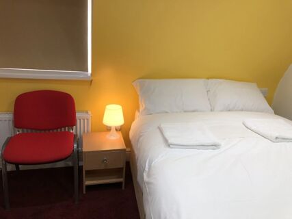 White bed with two pillows and towels beside a wooden nightstand with a lit lamp and a red chair in Leman House room