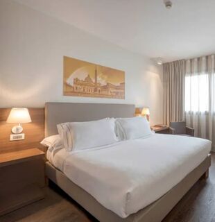 Bedroom at H10 Roma Citt&agrave;, Rom with a large bed, white linens, bedside lamps, a chair, wooden floor, and wall art above the bed