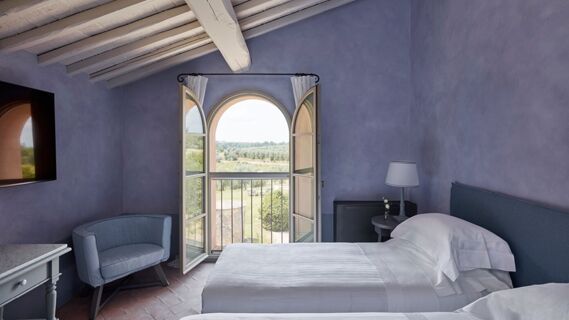 Bedroom with two white beds, blue walls and armchair, open arched window overlooking greenery at Podere San Filippo