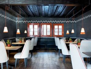 A dining area at COMO Alpina Dolomites featuring white cushioned chairs, wooden floors, wooden ceiling beams, and hanging pendant lights