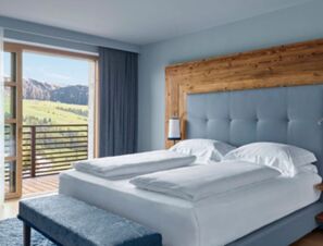 A hotel bedroom at COMO Alpina Dolomites with a large bed, blue padded headboard, bench, and mountain view balcony