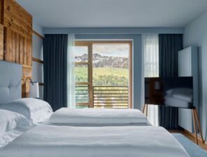 Hotel room at COMO Alpina Dolomites with two white beds, wooden wall panel, large window, and mountain view outside