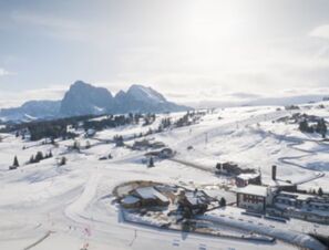 Snow-covered ski slopes and buildings at COMO Alpina Dolomites with rugged mountains in the background under a cloudy sky