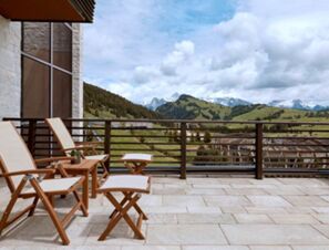 Terrace at COMO Alpina Dolomites with wooden chairs, small table, stone floor, and a railing overlooking green hills and mountains