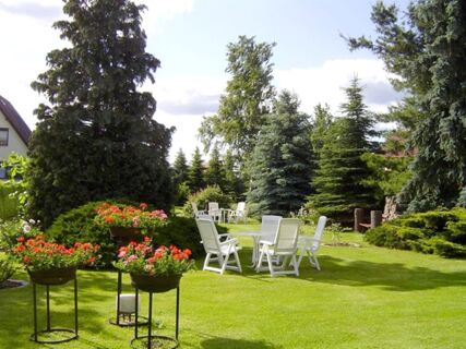 Green garden with white chairs and tables, flowering plants, and tall pine trees at Privatpension Flemminger Hof