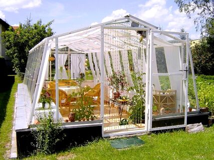 Greenhouse with white frame and glass walls featuring plants and wicker furniture at Privatpension Flemminger Hof in a garden setting
