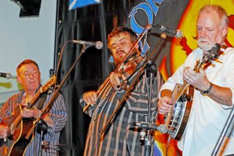 Drei Musiker spielen live Gitarre, Geige und Banjo auf einer B&uuml;hne im Sandgut Mihla Gasthof und Hotel bei einem Konzert.