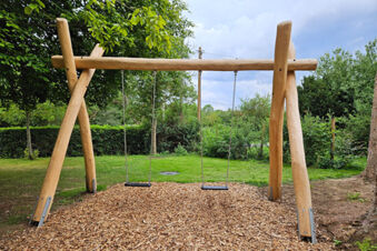 Zwei Holzschaukeln auf einem Mulchboden vor gr&uuml;nen B&auml;umen und Str&auml;uchern im Au&szlig;enbereich des Sandgut Mihla Gasthof und Hotel.