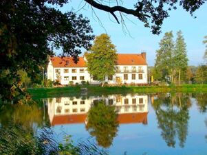 Hotel Zu den Schildb&uuml;rgern building with red roof and trees reflected in a calm pond under clear blue sky surrounded by green foliage