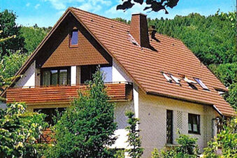 A two-story house with a brown tiled roof, white brick walls, a wooden balcony, and greenery surrounding it at Pension Haus Wiehenperle