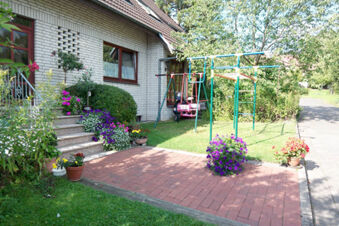 Green lawn and flowerpots surround a playground swing and climbing frame beside the brick house of Pension Haus Wiehenperle