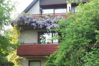 Woman wearing green dress standing on brown wooden balcony of Pension Haus Wiehenperle partially covered with purple flowers and surrounded by trees
