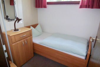 Small single wooden bed with light blue bedding, wooden cabinet with lamp, and window with red curtains in Pension Haus Wiehenperle