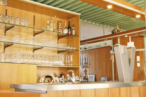 Bar area with glass shelves holding bottles and glasses, a metal counter with decorative items at Hotel Restaurant Illemann