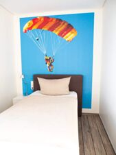 Kleines Einzelzimmer mit Holzboden und einem Bett, &uuml;ber dem an der Wand ein Fallschirmspringer auf blauem Hintergrund im JU 52 Hotel Arnsberg zu sehen ist.