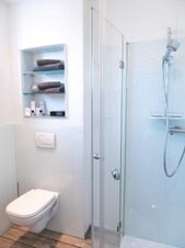Helles, modernes Badezimmer im JU 52 Hotel Arnsberg mit Dusche, WC und einem Regal mit Handt&uuml;chern und Pflegeprodukten.