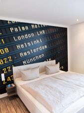 Hell eingerichtetes Hotelzimmer mit Doppelbett und Wandbild einer Anzeigetafel, modernes Design im JU 52 Hotel Arnsberg.