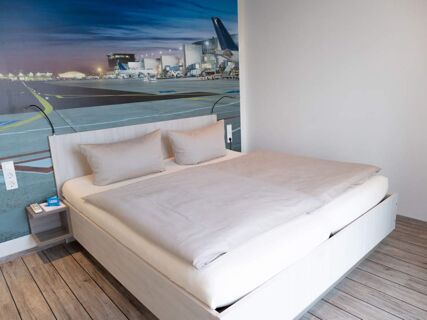 Modern eingerichtetes Hotelzimmer mit Doppelbett und Flughafenwandbild im JU 52 Hotel Arnsberg, schlicht und einladend gestaltet.