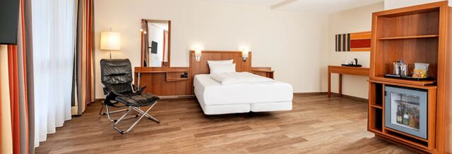 Room interior at NH Hotel M&uuml;nchen Dornach featuring a single bed, black chair, wooden furniture, and a minibar fridge visible on wooden flooring