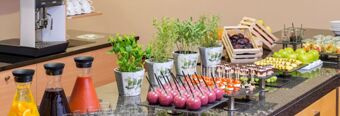 A buffet table at NH Hotel M&uuml;nchen Dornach displaying juices, potted herbs, fruit baskets, and assorted desserts and appetizers