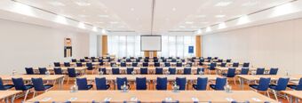 Large conference room at NH Hotel M&uuml;nchen Dornach with rows of tables and blue chairs facing a projector screen and windows