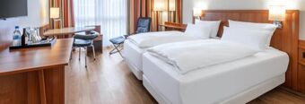 Two single beds with white linens, wooden headboard, desk with chair, and window with curtains in NH Hotel M&uuml;nchen Dornach room