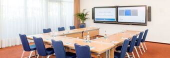 Conference room at NH Hotel M&uuml;nchen Dornach with U-shaped tables, blue chairs, water bottles, glasses, and dual screens on wall