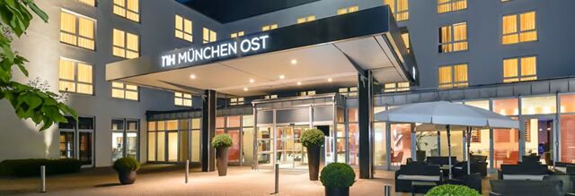 Entrance of NH Hotel M&uuml;nchen Dornach with illuminated canopy, glass doors, and outdoor seating area with umbrella at night