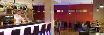 A bartender pouring a drink behind a lit bar counter with stools and seating area in NH Hotel M&uuml;nchen Dornach