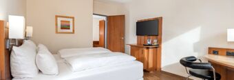 Bright hotel room at NH Hotel M&uuml;nchen Dornach with a bed, wooden furniture, mounted TV, desk, chair, and wall art